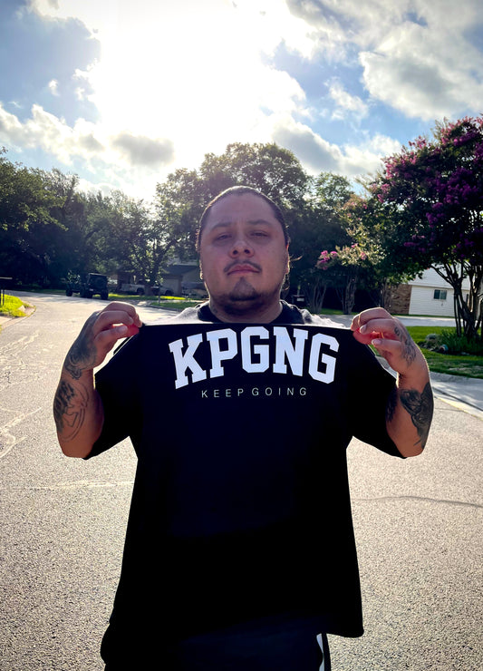 Person wearing a black t-shirt with 'KPGNG KEEP GOING' text outdoors on a sunny day.