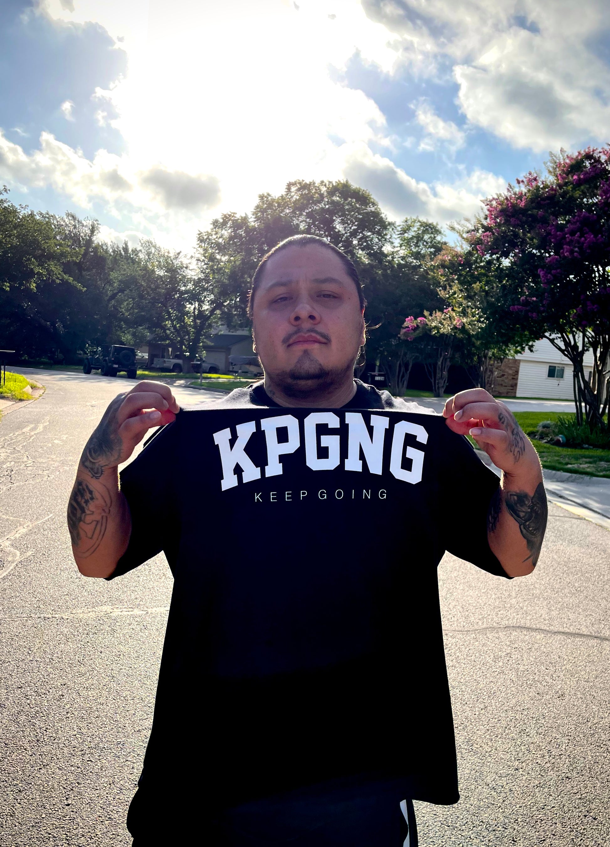 Person wearing a black t-shirt with 'KPGNG KEEP GOING' text outdoors on a sunny day.