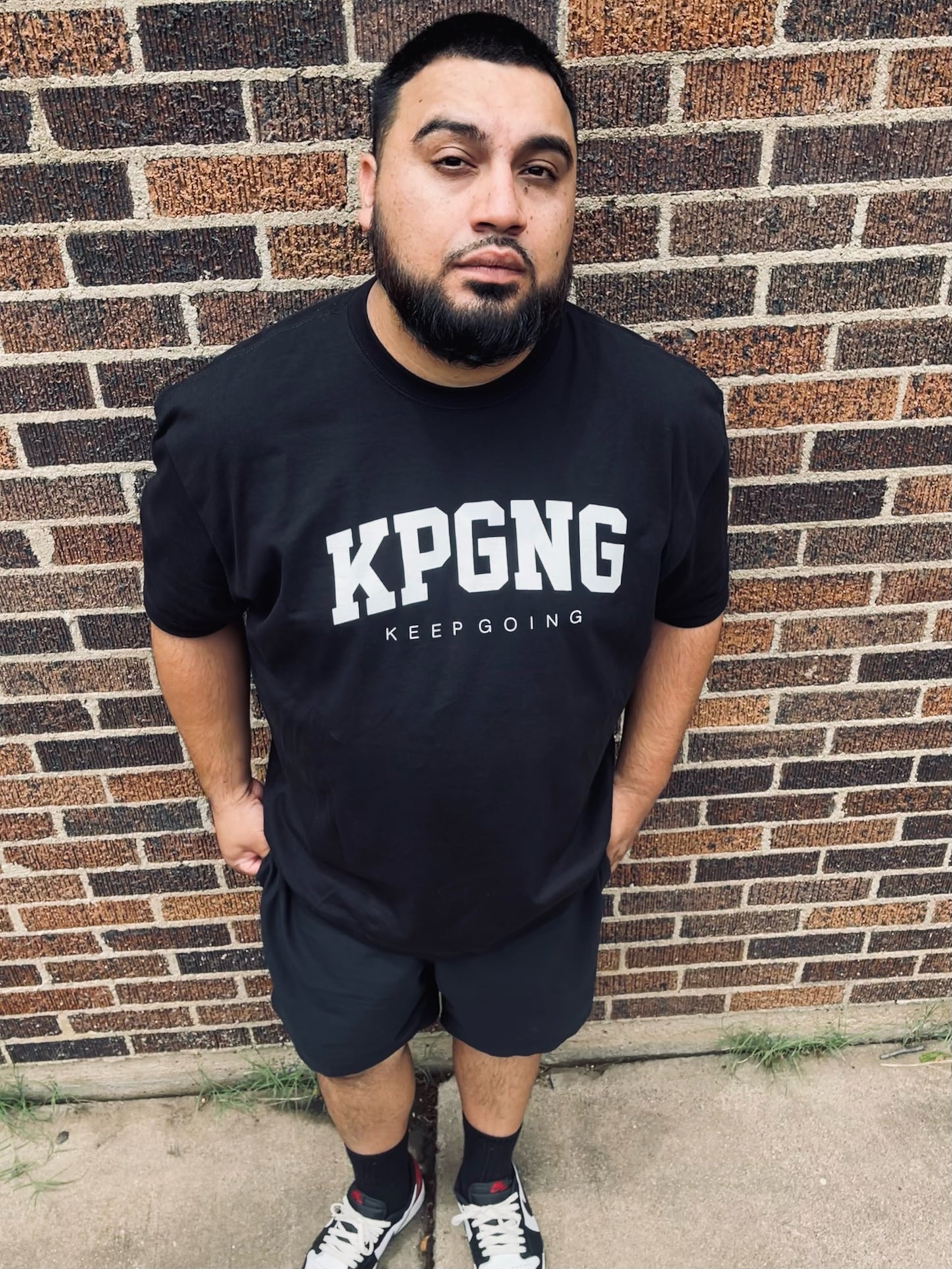 Man wearing a black t-shirt with 'KPGNG KEEP GOING' text standing against a brick wall.