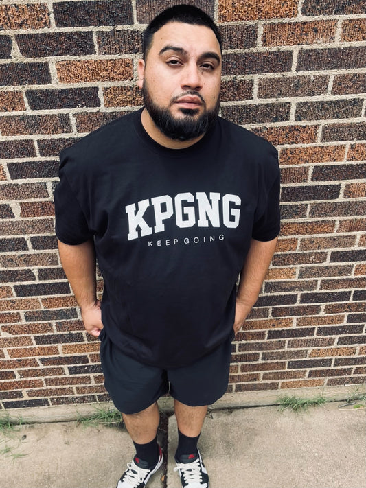 Man wearing a black t-shirt with 'KPGNG KEEP GOING' text standing against a brick wall.